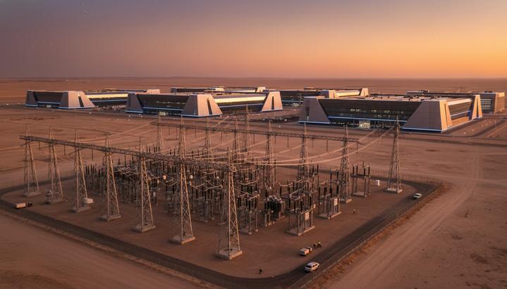 Огромный современный дата-центр на фоне заката, с мощной электроподстанцией и линиями электропередачи на переднем плане.